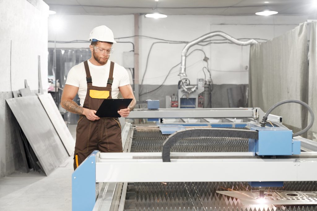 Front view of professional male mechanic standing near cnc and controlling process of plasma cutting on factory. Enginner in protective glasses and helmet keeping folder and reading. Concept of metal.